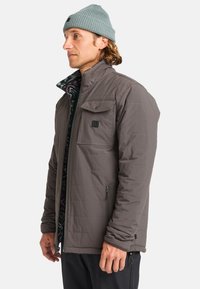 Chaqueta aislante gris con bolsillos con cremallera, cuello alzado y puños ajustables. Presenta un forro con pattern debajo del cuello.
