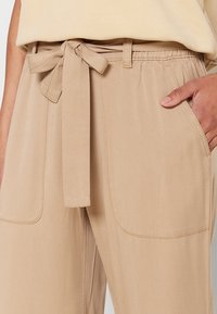 Pantalon beige taille haute avec poches plaquées à l’avant et ceinture en tissu nouée, porté avec un top beige assorti, main dans la poche.
