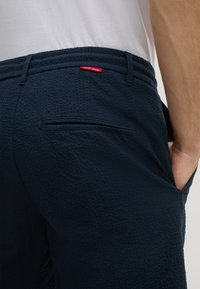 Pantaloni corti blu navy in tessuto testurizzato, con una tasca posteriore e dettaglio etichetta rossa. Vita piatta con cucitura orizzontale.