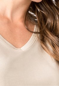 Beżowy top z dekoltem w serek o gładkiej, miękkiej fakturze. Posiada krótki dekolt i lekki połysk. Podkreślony przez luźne, falujące włosy w pobliżu.