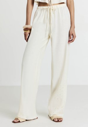 Pantalons à jambes larges en tissu crème doux et texturé, avec une taille élastique et des accents floraux brodés près de l'ourlet.