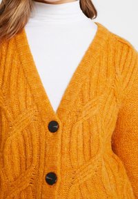 Cardigan lavorato a maglia arancione con scollatura a V, caratterizzato da un motivo a torsione testurizzato, due bottoni neri e una finitura morbida e pelosa.
