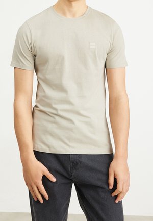 Mężczyzna ubrany w beżowy t-shirt z krótkim rękawem i okrągłym dekoltem oraz czarne spodnie, stojący na tle jasnego, jednolitego tła.