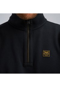 Crni pulover s polu-zip zatvaračem izrađen od mekog materijala, s zlatnom prugom na zatvaraču i pravokutnom oznakom branda na lijevoj strani prsnog koša.