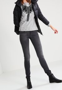Giacca a vento nera, maglione grigio con design ricamato in nero, jeans scuri aderenti, stivaletti neri, in piedi su uno sfondo bianco.