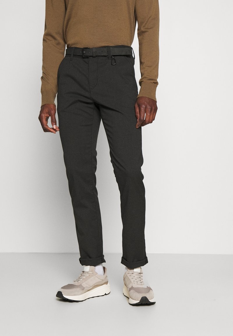 Tummanharmaat räätälöidyt housut slim fit -leikkauksella, joissa on vyö, yhdistetty ruskeaan neuleeseen ja vaaleisiin lenkkarikenkiin, yksinkertaisella taustalla.