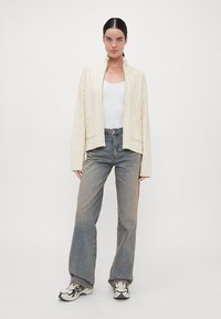 Λευκό πλεχτό καζί που έχει ψηλό γιακά και σχέδιο καλωδίου, συνδυασμένο με ανοιχτό μπλε τζιν κατευθείαν πόδια και μαύρα-άσπρα αθλητικά παπούτσια.