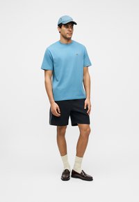 Mann trägt hellblauen Hut und T-Shirt, schwarze Shorts, beige Socken und dunkelbraune Mokassins, steht vor einfarbigem Hintergrund.