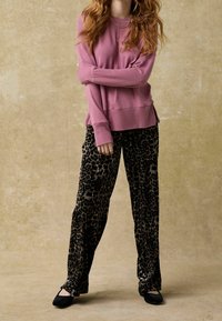 Różowy sweter o luźnym kroju, z prążkowanymi mankietami, oraz aksamitne spodnie w panterkę, połączone z czarnymi balerinami. Prosty, warstwowy naszyjnik jako akcent.