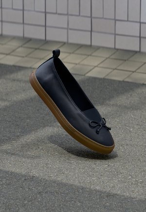 Sort læder slip-on sko med brun gummisål og bundet sløjfedetalje, svævende over en grå fortov ved siden af en flisebelagt væg.