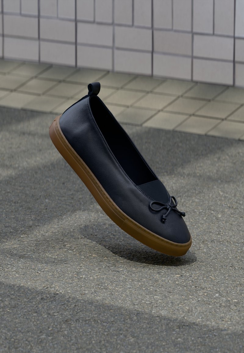 Μαύρο δερμάτινο παπούτσι χωρίς κορδόνια με καφέ λαστιχένια σόλα και δεμένη λεπτομέρεια φιόγκου, αιωρούμενο πάνω από γκρι πεζοδρόμιο δίπλα σε πλακιδωτό τοίχο.