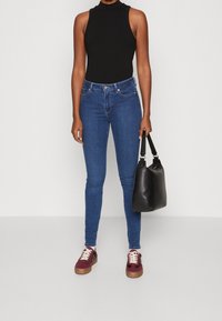 Top sin mangas negro de canalé, jeans ajustados azules y zapatillas bajas burdeos. Sosteniendo un bolso negro con asas cortas.
