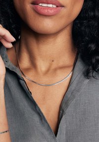 Srebrny metaliczny naszyjnik o gładkim, zakrzywionym kształcie, noszony na nagiej szyi, w połączeniu z ciemnoszarym t-shirtem. Widoczny naturalny odcień skóry.