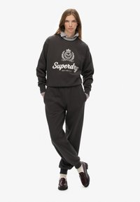 Vrouw in een zwart Superdry sweatshirt en bijpassende joggers, gelaagd over een gestreept shirt, met witte sokken en donkere loafers, staande met gekruiste benen.
