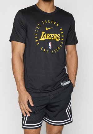 Mand iført en sort Los Angeles Lakers T-shirt og sorte basketballshorts med hvide striber, står foran en ensfarvet baggrund.