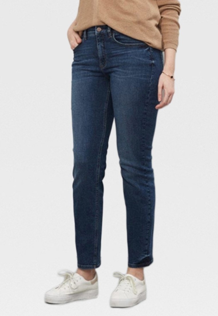 Persona vestida con jeans ajustados azul oscuro, zapatillas blancas y una camiseta de manga larga beige, de pie con una mano en el bolsillo.