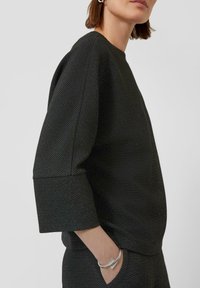 Zwart gestructureerd pullover met driekwart mouwen, voorzien van een ronde halslijn en een geribde zoom. Zilveren armband benadrukt de pols.