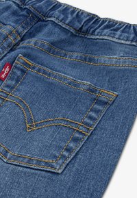 Μπλε υφασμάτινο τζιν με μεγάλη πίσω τσέπη που διαθέτει κίτρινα ράμματα και μια μικρή ετικέτα με το λογότυπο κοντά στη μέση. Απαλή, ανθεκτική υφή.