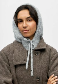 Abrigo de lana marrón con un acabado texturizado, que cuenta con una capucha de punto gris atada en el cuello con lazos, creando un diseño en capas.