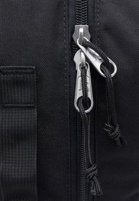 Sac en tissu noir avec fermeture éclair, équipé de tirettes à double zip en métal argenté et d'une cordelière tressée noire. Surface texturée et design robuste.