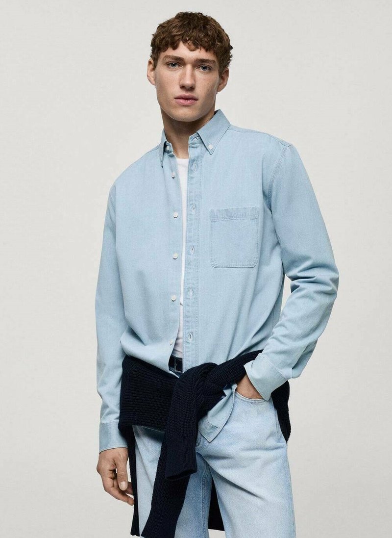 Camicia in denim azzurro chiaro con bottoni e tasca, indossata sopra una t-shirt bianca, abbinata a jeans azzurri chiari e a un maglione blu navy legato in vita.