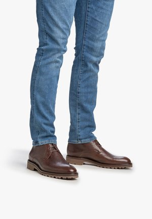 Botas de cuero marrón hasta el tobillo con un acabado texturizado, diseño de cordones y una suela robusta, combinadas con jeans ajustados azules.