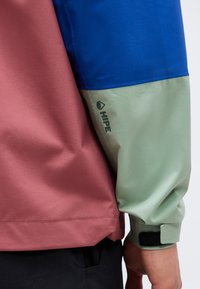 Μανίκι μπουφάν με μπλε και ανοιχτό πράσινα χρωματικά μπλοκ, λογότυπο "HIPE" στο μπράτσο και ρυθμιζόμενο μανσέτα με βέλκρο δίπλα στο σώμα ενός μπορντό μπουφάν.