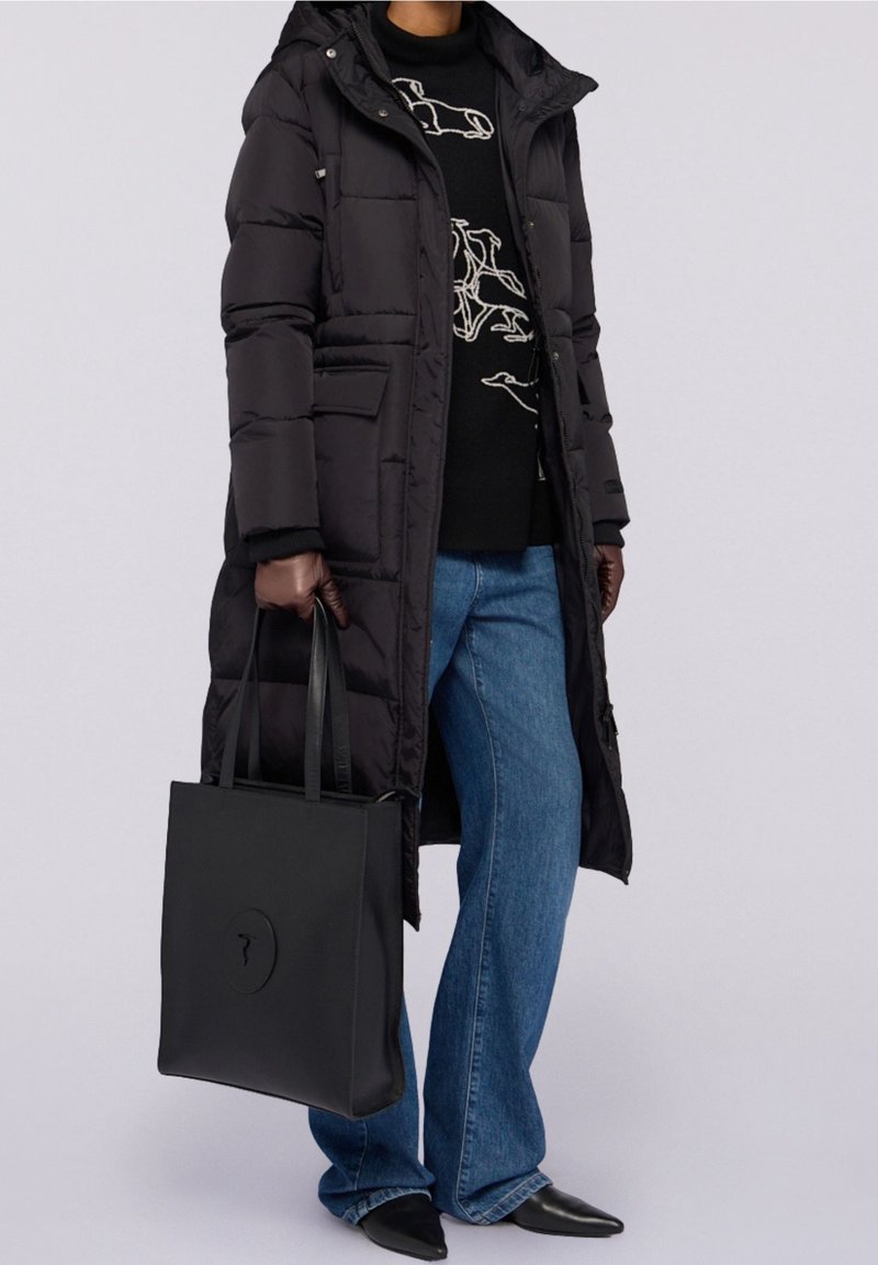 Personne en long manteau matelassé noir, pull noir avec un dessin linéaire blanc, jean bleu, gants noirs, tenant un grand sac à main noir.