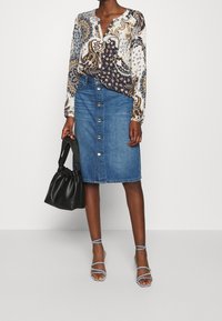 Blusa a maniche lunghe con motivo paisley in blu, beige e bianco, abbinata a una gonna in denim azzurro a lunghezza ginocchio con dettagli a bottone. Borsa nera.