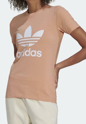 Osoba nosząca brzoskwiniową koszulkę Adidas z dużym białym logo i jasne spodnie, stojąca z jedną ręką na biodrze.