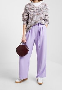 Pull en tricot en fil multicolore, associé à un pantalon lavande taille haute et des baskets blanches. Tenant un sac à main rond bordeaux.