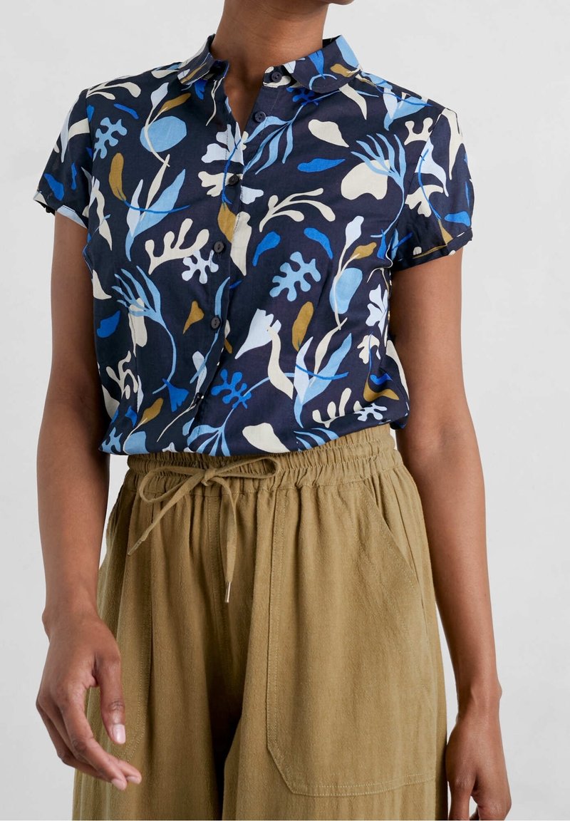 Kvinna i en kortärmad marinblå skjorta med blå, vita och beige blommönster, nedstoppad i högmidjade beige byxor med snöre.