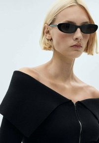 Zwarte, off-the-shoulder gebreide top met een rits, voorzien van een brede kraag. Het model draagt stijlvolle zwarte zonnebril tegen een egale achtergrond.