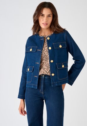 Femme portant une veste en jean bleu foncé avec des boutons dorés sur un haut imprimé léopard et un jean en jean bleu foncé assorti, debout avec la main dans la poche.