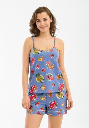 Giovane donna che indossa un top blu con spalline sottili e pantaloncini con motivi colorati a frutti tropicali, sorridente con le mani rilassate ai lati.