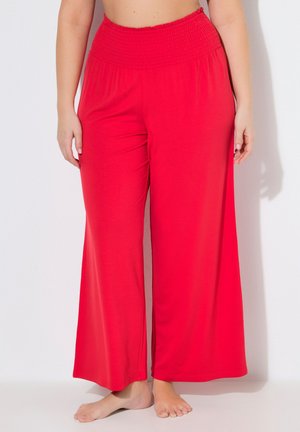 Frau trägt weite, knallrote, hoch taillierte Hosen mit weitem Bein, steht barfuß vor weißem Hintergrund.
