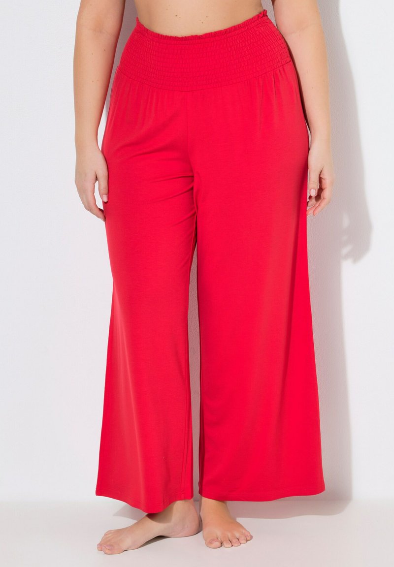 Kvinde iført løstsiddende, knaldrøde bukser med høj talje og vide ben, stående barfodet mod en hvid baggrund.