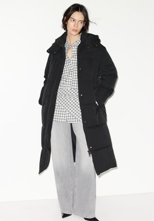 Geacă lungă matlasată neagră, cu glugă, prevăzută cu fermoar și capse, purtată peste o cămașă gri în carouri și pantaloni largi de culoare gri deschis.