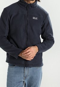 Marinblå fleecejacka med hög krage, hela dragkedjan och logotyp på vänster bröst. Den har långa ärmar och en avslappnad passform.