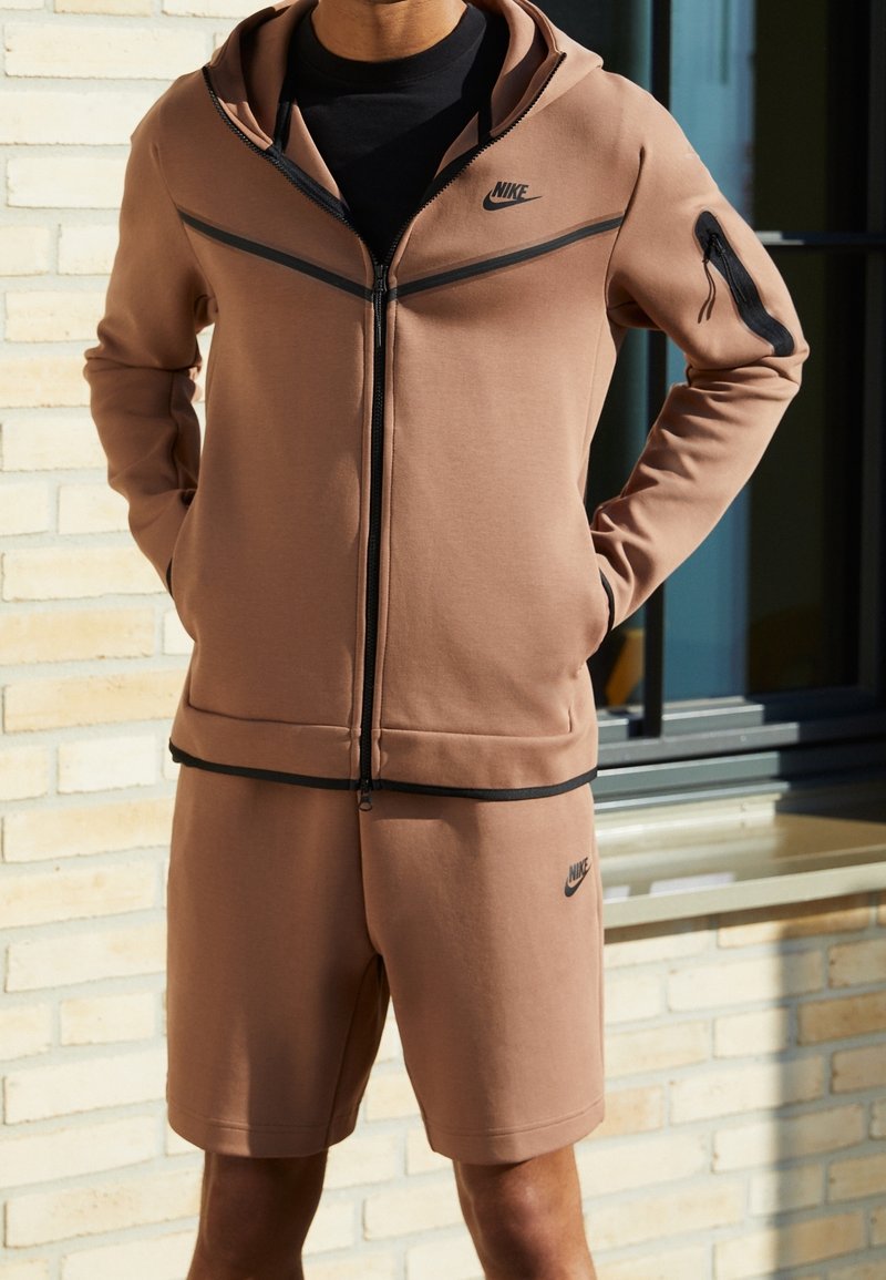 Sada mikiny na zip a šortek Nike v tlumené hnědé barvě. Obsahuje černé akcenty a logo, s hladkou texturou a přiléhavým střihem.