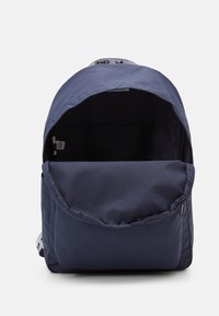 Navy blue fabric backpack with a rounded top, flap closure, front pocket, and a grey logo tag on the top interior edge.