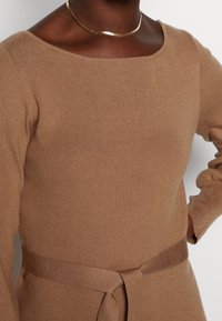 Lähikuva henkilöstä, joka pukee päällään ruskean neulotun pitkähihasen mekon, johon kuuluu samasta langasta tehty vyö ja ohut kullanvärinen kaulaketju, tasaisella taustalla.