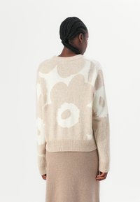 Stickad tröja med en ljusbeige bas som har stora vita blommönster, ribbad kant och en avslappnad passform. Slät textur.