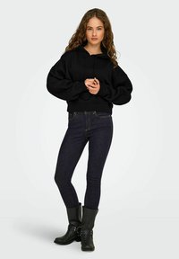 Felpa nera corta con cappuccio regolabile, abbinata a jeans in denim scuro. Modella che indossa stivali neri alla caviglia con fibbie.
