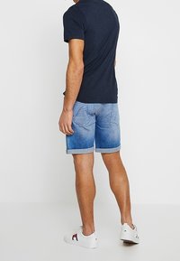 Hombre vestido con una camiseta azul marino, pantalones cortos de denim claro enrollados y zapatillas blancas, de pie contra un fondo blanco liso.