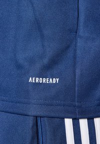 Blauwe sportstof met een gestructureerd oppervlak, met het "AEROREADY" logo in het wit. Bevat een ritsdetail en contrasterende witte strepen.