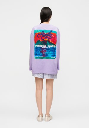 Frau trägt einen lavendelfarbenen Oversize-Pullover mit buntem „Paradise Island“-Motiv von Bergen und Meer auf dem Rücken, kombiniert mit karierten Shorts.