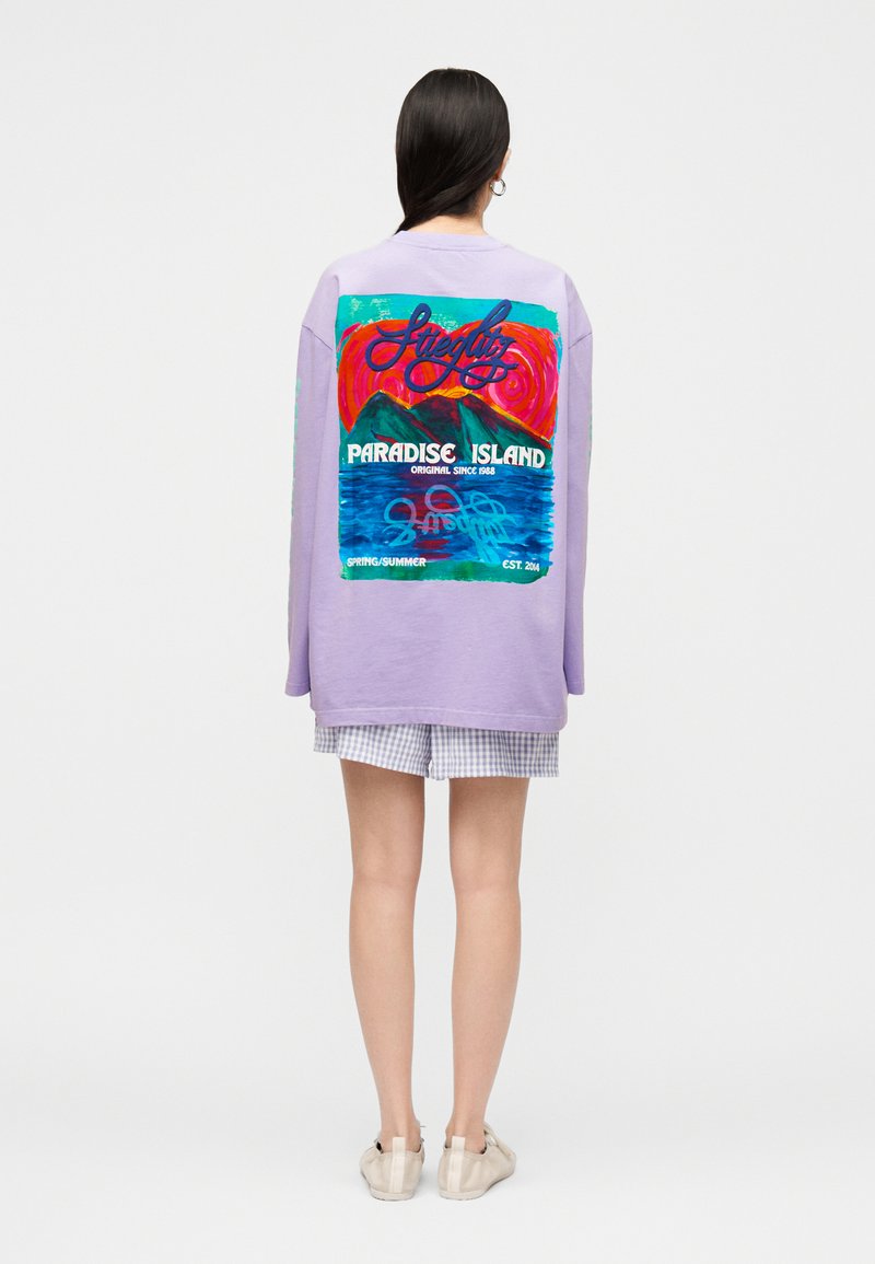 Γυναίκα που φοράει ένα λιλά φαρδύ φούτερ με πολύχρωμο γραφικό "Paradise Island" με βουνά και ωκεανό στην πλάτη, συνδυασμένο με καρό σορτς.