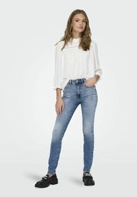 Blusa blanca de manga larga con cuello volante y detalles suavemente fruncidos, combinada con jeans ajustados de un azul claro que presentan detalles desgastados.