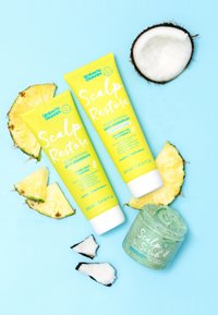 Geel tubes van "Scalp Restore" anti-roos shampoo en conditioner met ananasaccenten, plus een groene pot "Scalp Scrub", kokosstukjes, op een blauwe achtergrond.